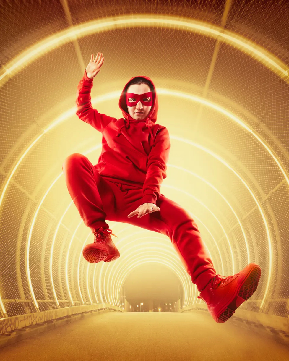 Pige i rødt tøj med rød hætte og rød maske springer op i luften i gul tunnel. Fra teaterforestilling Håndbog for superhelte på Odense Teater